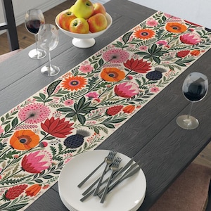 May include: A rectangular table runner featuring a vibrant botanical print with orange, red, and pink flowers, green foliage, and dark blue thistle accents on a cream background, laid across a dark wooden table with plates and wine glasses.