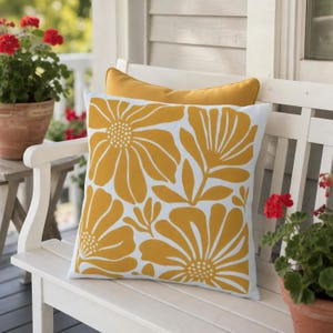 May include: A square throw pillow featuring a white background with a bold, mustard yellow floral pattern. The design consists of large, stylized flower heads and leaves arranged in a repeating graphic print. A solid mustard yellow pillow is visible behind.
