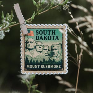 Puede incluir: Una pegatina con forma de sello postal con el Monte Rushmore, con el texto "SOUTH DAKOTA" y una bandera estadounidense. La pegatina está sujeta por una pinza de madera sobre un fondo natural borroso.