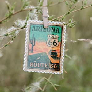 May include: A rectangular sticker shaped like a postage stamp featuring a desert landscape with a saguaro cactus, a road, and a car. The text reads Arizona and Route 66. The design uses orange, green, and black colors with a white scalloped border.