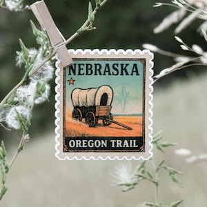 May include: A postage stamp design featuring a covered wagon on the Oregon Trail, with the text "NEBRASKA" and "OREGON TRAIL". The stamp is held by a wooden clothespin on a background of greenery.