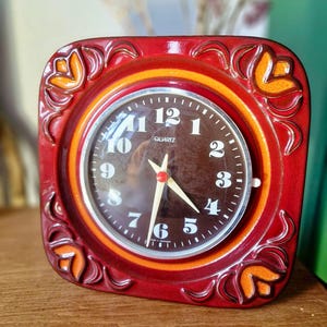 May include: A square ceramic quartz clock with a glossy red frame featuring embossed orange floral motifs in each corner. The circular clock face is dark brown with white Arabic numerals, a silver-tone bezel, and white hour and minute hands.