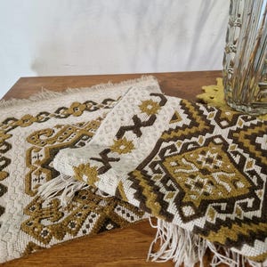 May include: Two rectangular woven placemats featuring a geometric pattern in shades of mustard yellow, dark brown, and cream. The fabric has a textured weave with fringed edges on the short sides, displaying a repeating diamond and cross motif.