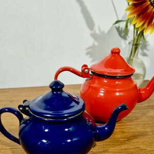 Op de afbeelding: Twee kleine, glanzende emaille theepotten met deksels en gebogen tuiten. De ene theepot is diep marineblauw, terwijl de andere fel oranje is. Beide hebben een ronde vorm, een scharnierend deksel met een knop en een handvat aan de zijkant.