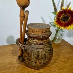 May include: A brown speckled ceramic pot with a cork lid and a side handle holding two wooden utensils. One utensil features a round head with small holes, while the other is a simple stick. The pot has a textured, earthy finish and a wide rim.