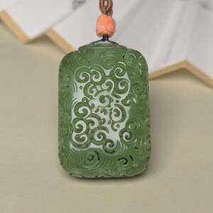 May include: A green jade pendant with detailed floral carvings. The rectangular pendant, in a light green shade, hangs from a brown cord with a peach bead. The design showcases swirling, symmetrical patterns, typical of jade craftsmanship.