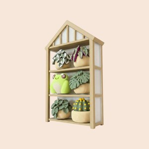 May include: A light brown wooden house-shaped shelf with three tiers. The top shelf holds a white and green patterned plant and a purple and green plant. The middle shelf features a light green frog figurine and a green leaf plant. The bottom shelf displays a green leafy plant and a round green cactus with yellow dots.