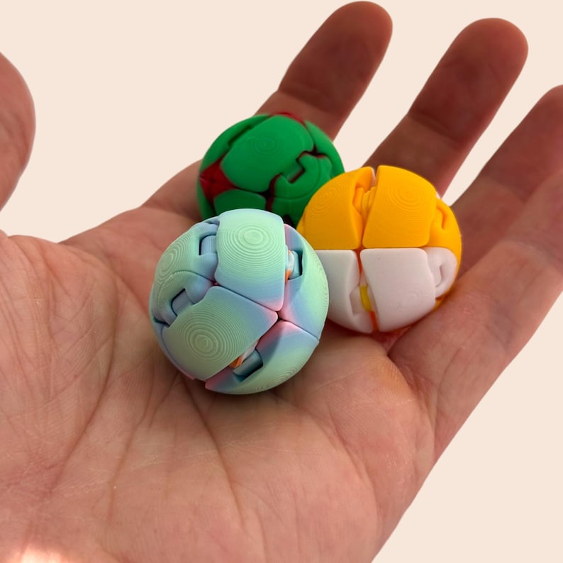 Pu&ograve; includere: Tre giocattoli puzzle sferici colorati in una mano. Uno &egrave; verde con dettagli rossi, un altro giallo e bianco, e il terzo blu e rosa. I giocattoli sono fatti di pezzi ad incastro. Ogni giocattolo misura circa 5 cm di diametro.