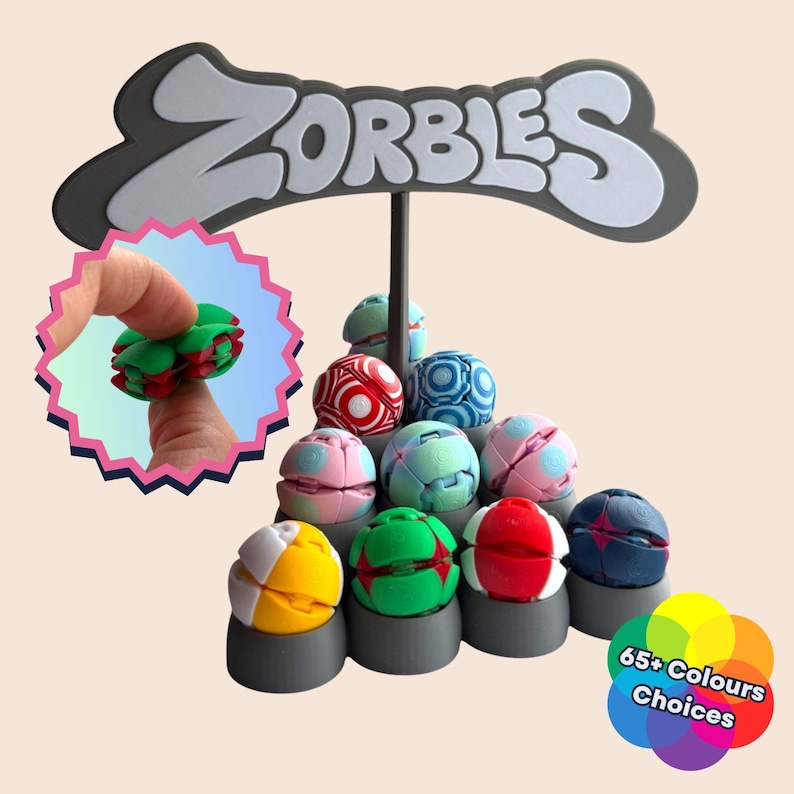 Pu&ograve; includere: Un'esposizione di Zorbies colorati, giocattoli puzzle sferici, con un cartello che dice "Zorbies". I giocattoli sono in vari colori, tra cui rosso, verde, blu e rosa. Un'immagine pi&ugrave; piccola mostra un giocattolo Zorbies tenuto in mano. L'immagine include anche il testo "65+ Colours Choices".