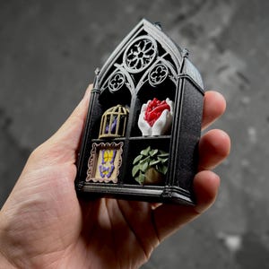 May include: A black 3D-printed miniature gothic window frame featuring four compartments. The top left holds a gold birdcage, the top right contains white hands cradling a red heart, the bottom left shows a framed yellow butterfly, and the bottom right displays a green potted plant.