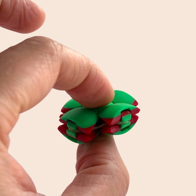 Pu&ograve; includere: Una palla puzzle geometrica verde e rossa, tenuta tra le dita. &Egrave; composta da pezzi ad incastro, con sezioni verdi e accenti rossi. Il design &egrave; intricato e visivamente accattivante, suggerendo un rompicapo o un oggetto decorativo.