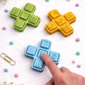 May include: Three colourful, cross-shaped sensory toys with textured surfaces. One is green, one is yellow, and one is blue. A finger presses a button on the blue toy. The toys are surrounded by small, decorative beads and flowers.