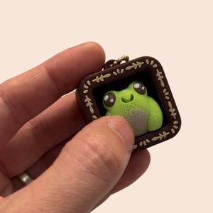 May include: A small, square, dark brown keychain frame featuring a light green frog with large, dark eyes and a pale belly. The frame is decorated with a repeating cream-coloured leaf pattern along its edges and includes a gold-toned metal chain.