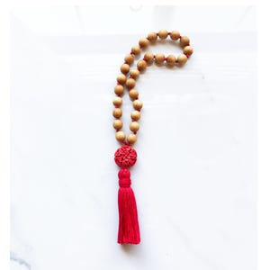 May include: A wooden bead necklace with a red tassel and a carved red accent piece. The beads are light brown and strung on a red cord. The tassel is a vibrant red color.