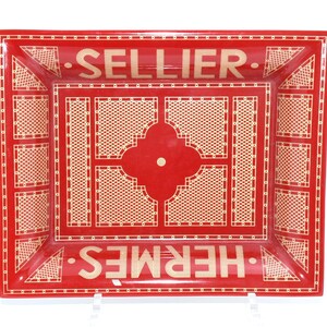 May include: A rectangular red tray with a cream-colored lattice design and the words "SELLIER" and "HERMES" in cream-colored text. The tray has a raised edge and a decorative center.
