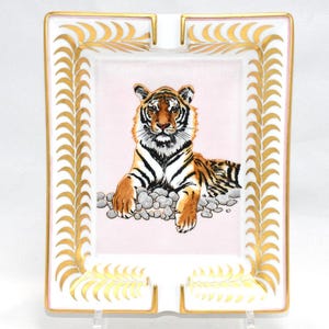 May include: Rectangular ashtray with a pink center featuring a tiger illustration. The ashtray has a white border with gold leaf accents and gold trim. The tiger is orange, black, and white, resting on gray stones.