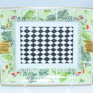 May include: Rectangular ceramic ashtray with a gold rim and accents. The ashtray features a central black and white checkered pattern, surrounded by a light green border with tropical leaf and floral designs. Two gold-colored rests are on the sides.
