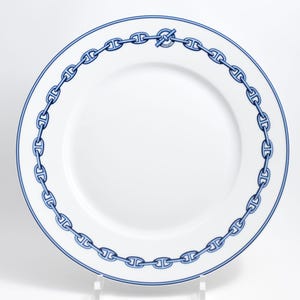 May include: White ceramic plate with a blue chain link design around the rim. The plate has a blue border and a simple, elegant aesthetic. The chain link pattern is a repeating design.