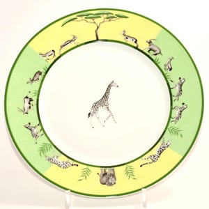 May include: A decorative plate with a safari theme. The plate features a giraffe in the center, surrounded by illustrations of zebras, gazelles, and a lion. The rim has a green border with a yellow and green background.