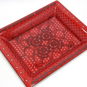 May include: A square, red ceramic tray with a glossy finish. The tray features a paisley border and a central design of interconnected circles and dots. The background is covered in white polka dots.