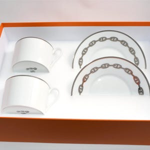 May include: A boxed set of two white teacups and two saucers. The cups and saucers have a dark gray trim and a chain design. The box is orange with a white interior, presenting the set in a neat arrangement.
