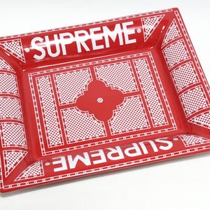 May include: A red rectangular tray with white geometric patterns and the word "SUPREME" in white block letters. The tray has a raised edge and a decorative floral design in the center.