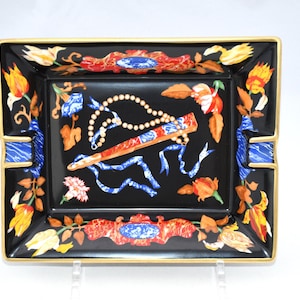 May include: Black rectangular ashtray with a gold rim and floral design. The ashtray features a central design of a fan, pearls, and ribbons. The corners have blue accents.