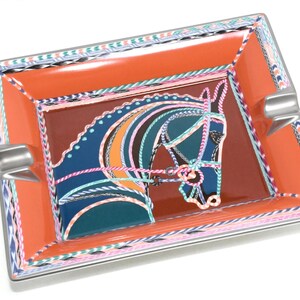 May include: Rectangular orange ashtray with a horse head design. The horse head is depicted in blue, teal, and pink with a brown background. The ashtray has a silver rim and two cigarette rests.
