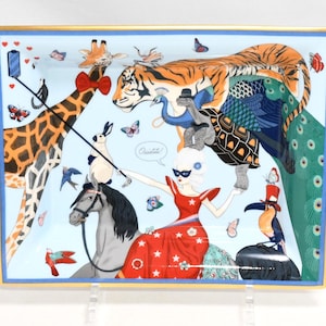 May include: Rectangular decorative tray with a light blue background and a gold border. The tray features a whimsical illustration of animals, including a giraffe, tiger, peacock, and toucan, with a woman in a red dress. Butterflies and birds are also depicted.
