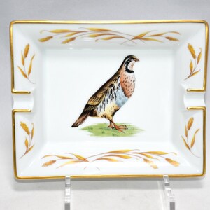 May include: Rectangular white porcelain ashtray with a gold rim and corner accents. The center features a detailed illustration of a bird standing on green grass. The ashtray has four cigarette rests.