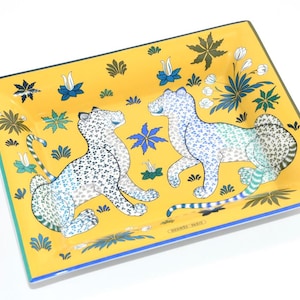 May include: Rectangular decorative tray with a yellow background and a blue and green border. The tray features two stylized snow leopards with blue and white spots, surrounded by floral and leaf motifs. The words "HERMES PARIS" are printed on the tray.