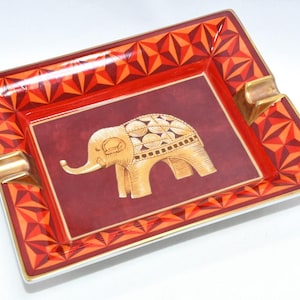 May include: Rectangular ashtray with a geometric design in shades of red and orange. The center features a gold-colored elephant illustration on a burgundy background. The ashtray has gold-colored rests on each side.