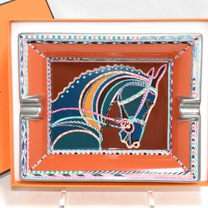 May include: Rectangular orange ashtray with a horse head design in blue, orange, and pink. The ashtray has a brown center and silver rests. The border features a colorful geometric pattern. The ashtray is in an orange box with the Hermes logo.