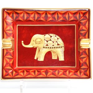 May include: Rectangular ashtray with a red background and a gold elephant design. The border features a geometric pattern in shades of red and orange, with gold accents. The ashtray has gold-colored rests.