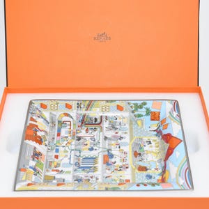 May include: A rectangular decorative tray with a colorful, detailed illustration of a city scene. The tray is presented inside an orange box with a white interior. The tray's design includes buildings, figures, and various architectural elements.