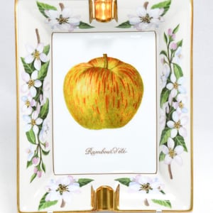May include: Rectangular porcelain ashtray with a gold rim and two gold rests. The center features a detailed illustration of a yellow and red apple with the text "Rambour d'été". White flowers and green leaves decorate the border.