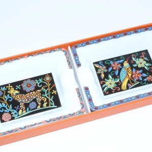 May include: A set of two decorative ceramic trays with a black background and colorful floral and animal designs. The trays are in a rectangular orange box with a white interior and a patterned border.