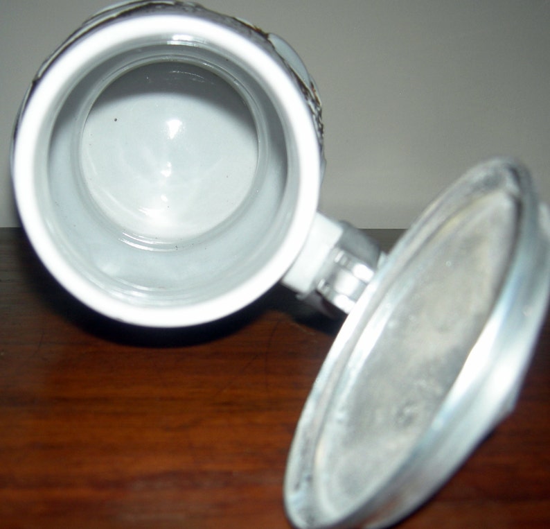 May include: A white ceramic beer stein with a metal lid. The stein has a traditional design with a raised handle and a round body. The lid is hinged and has a small knob on top.