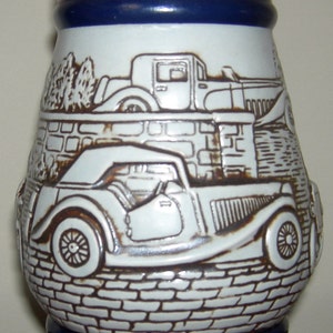 May include: A white ceramic beer stein with a silver lid, featuring a brown and white illustration of a 1936 MG and a 1927 Bugatti. The stein has a blue band around the middle and red and blue bands around the bottom, with the text "1936 MG - 1927 Bugatti" printed on the bottom.