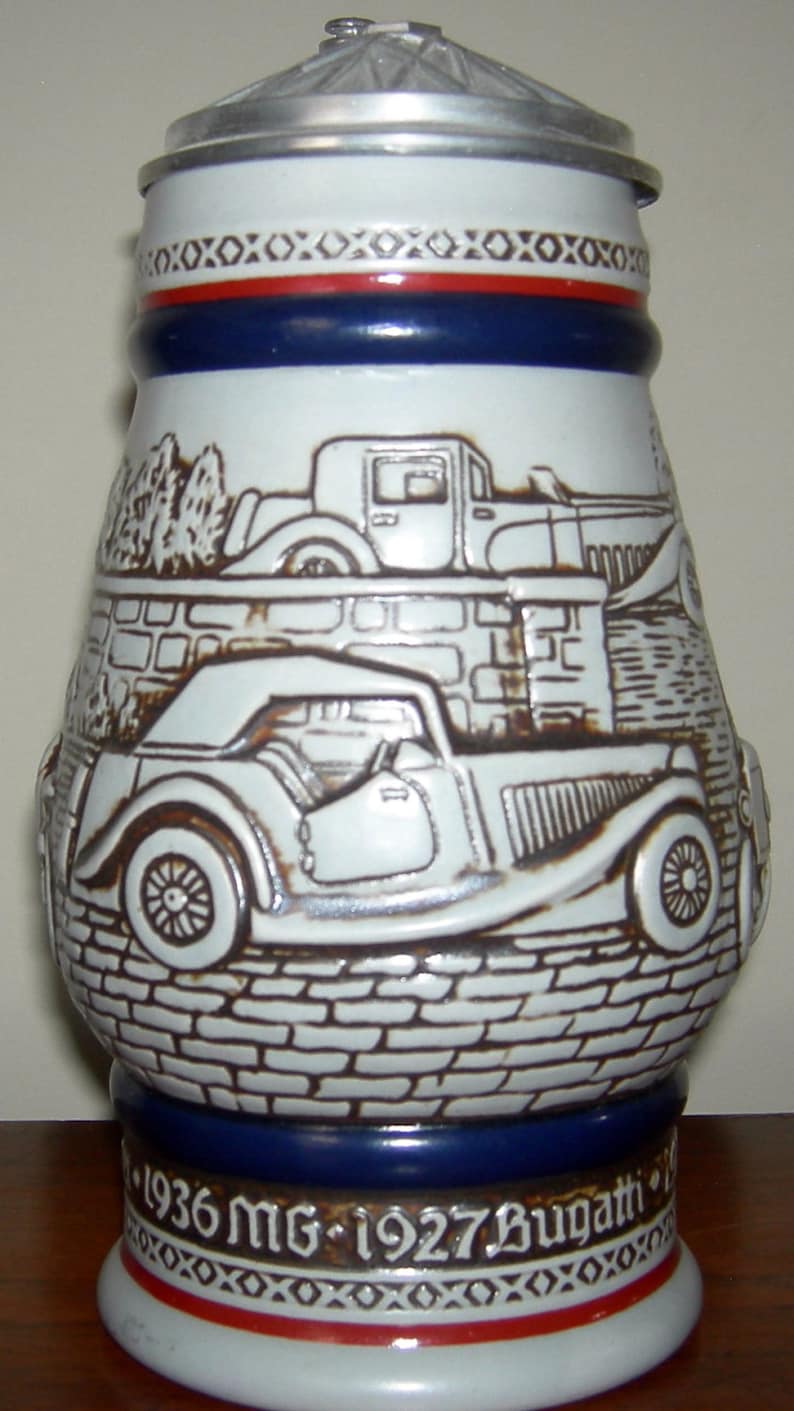 May include: A white ceramic beer stein with a silver lid, featuring a brown and white illustration of a 1936 MG and a 1927 Bugatti. The stein has a blue band around the middle and red and blue bands around the bottom, with the text "1936 MG - 1927 Bugatti" printed on the bottom.