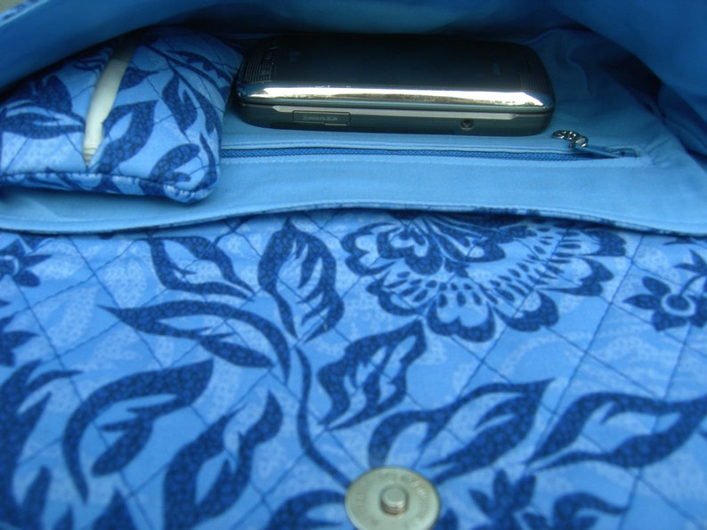 K&ouml;nnte beinhalten: Eine blaue gesteppte Handtasche mit Blumenmuster. Die Tasche hat eine Rei&szlig;verschlusstasche und ein Handyfach.
