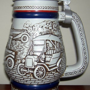 May include: A white ceramic beer stein with a blue and red band around the middle. The stein features a detailed image of a 1911 Ford Model T car. The stein has a silver lid and handle. The text "1911 Ford Model T" is printed on the base of the stein.