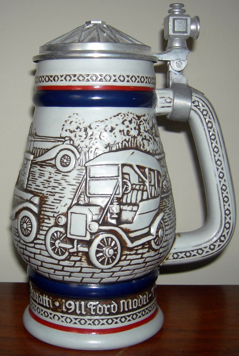 May include: A white ceramic beer stein with a blue and red band around the middle. The stein features a detailed image of a 1911 Ford Model T car. The stein has a silver lid and handle. The text "1911 Ford Model T" is printed on the base of the stein.