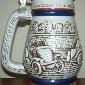 May include: A white ceramic beer stein with a brown and white image of a vintage car. The stein has a blue and red band around the top and the words "Stanley Steamer - 1936" on the bottom.