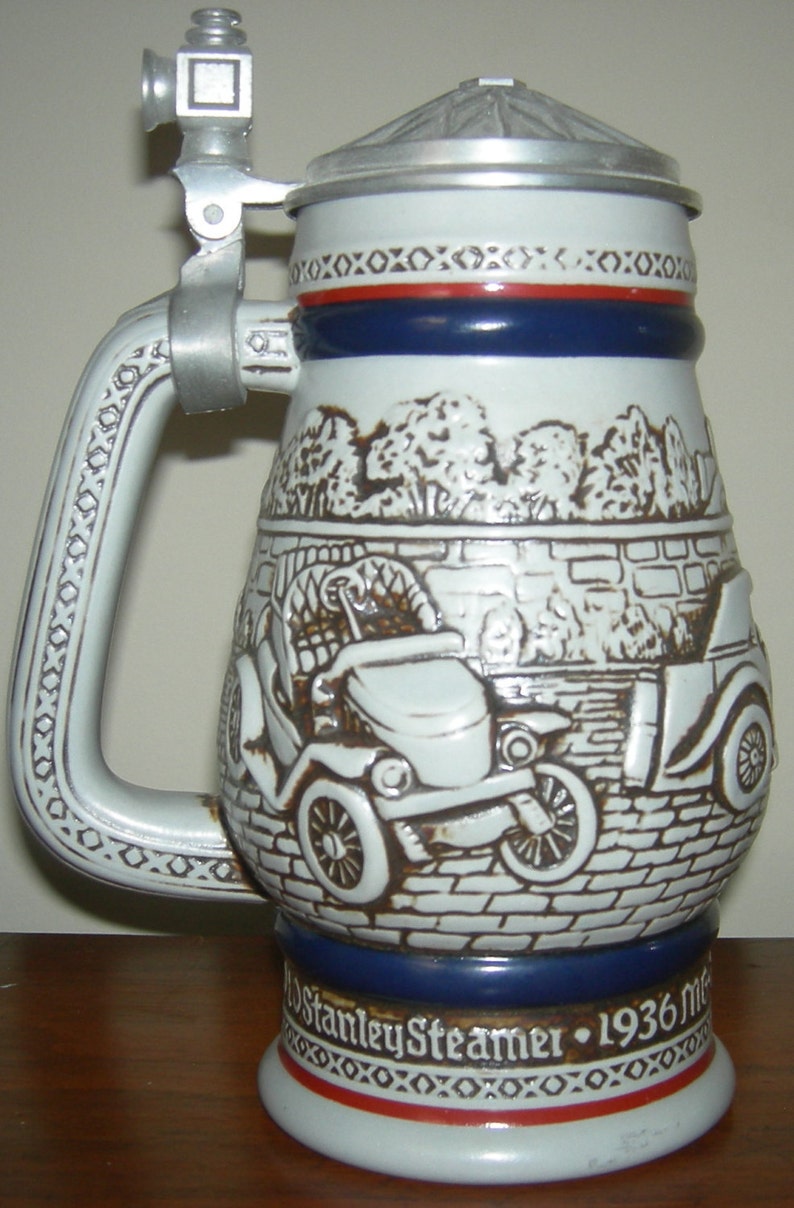May include: A white ceramic beer stein with a brown and white image of a vintage car. The stein has a blue and red band around the top and the words "Stanley Steamer - 1936" on the bottom.
