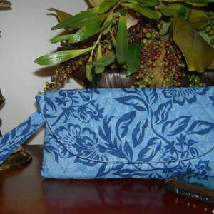 K&ouml;nnte beinhalten: Eine blaue gesteppte Clutch mit floralem Muster und einem passenden Riemen. Die Clutch hat einen Klappverschluss und ein kleines Fach auf der R&uuml;ckseite. Ein schwarzes Handy liegt auf einem Tisch neben der Clutch.