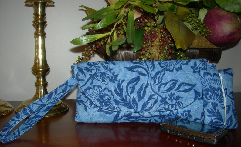 K&ouml;nnte beinhalten: Eine blaue gesteppte Clutch mit floralem Muster und einem passenden Riemen. Die Clutch hat einen Klappverschluss und ein kleines Fach auf der R&uuml;ckseite. Ein schwarzes Handy liegt auf einem Tisch neben der Clutch.
