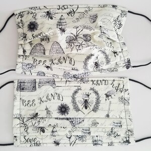 May include: Two white face masks with black elastic ear loops. The masks are patterned with black illustrations of bees, beehives, flowers, and script that reads "Bee Kind" and "Save the Bees".