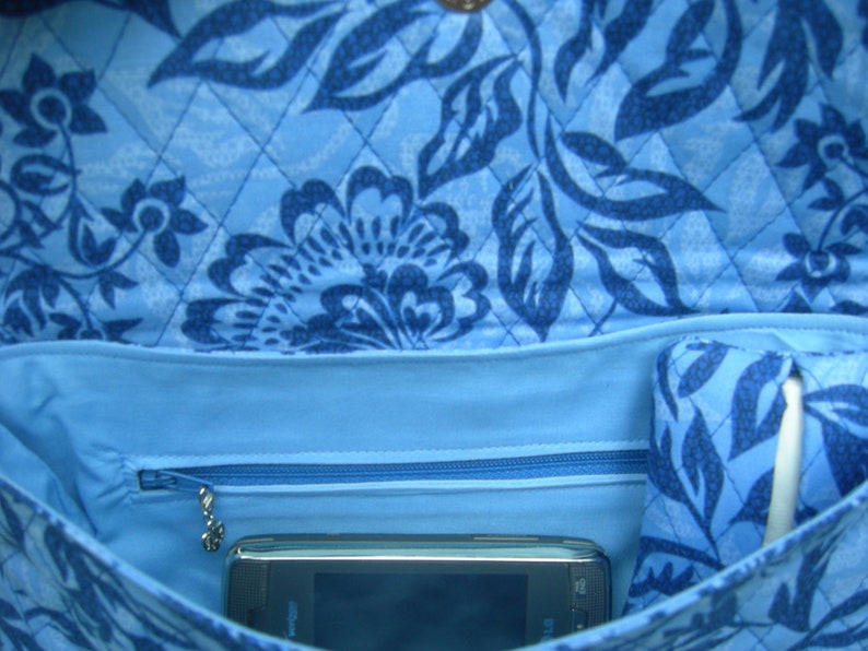 K&ouml;nnte beinhalten: Eine blaue gesteppte Tasche mit einem Blumenmuster. Die Tasche hat eine Rei&szlig;verschlusstasche und ein kleines Telefon im Inneren.