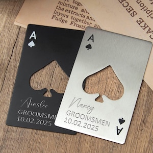 Pode incluir: Dois abridores de garrafa em forma de Ás de Espadas, um preto e outro prateado, personalizados com nomes e datas. Os abridores estão sobre uma superfície de madeira, com uma garrafa de Hennessy e uma receita antiga no fundo.